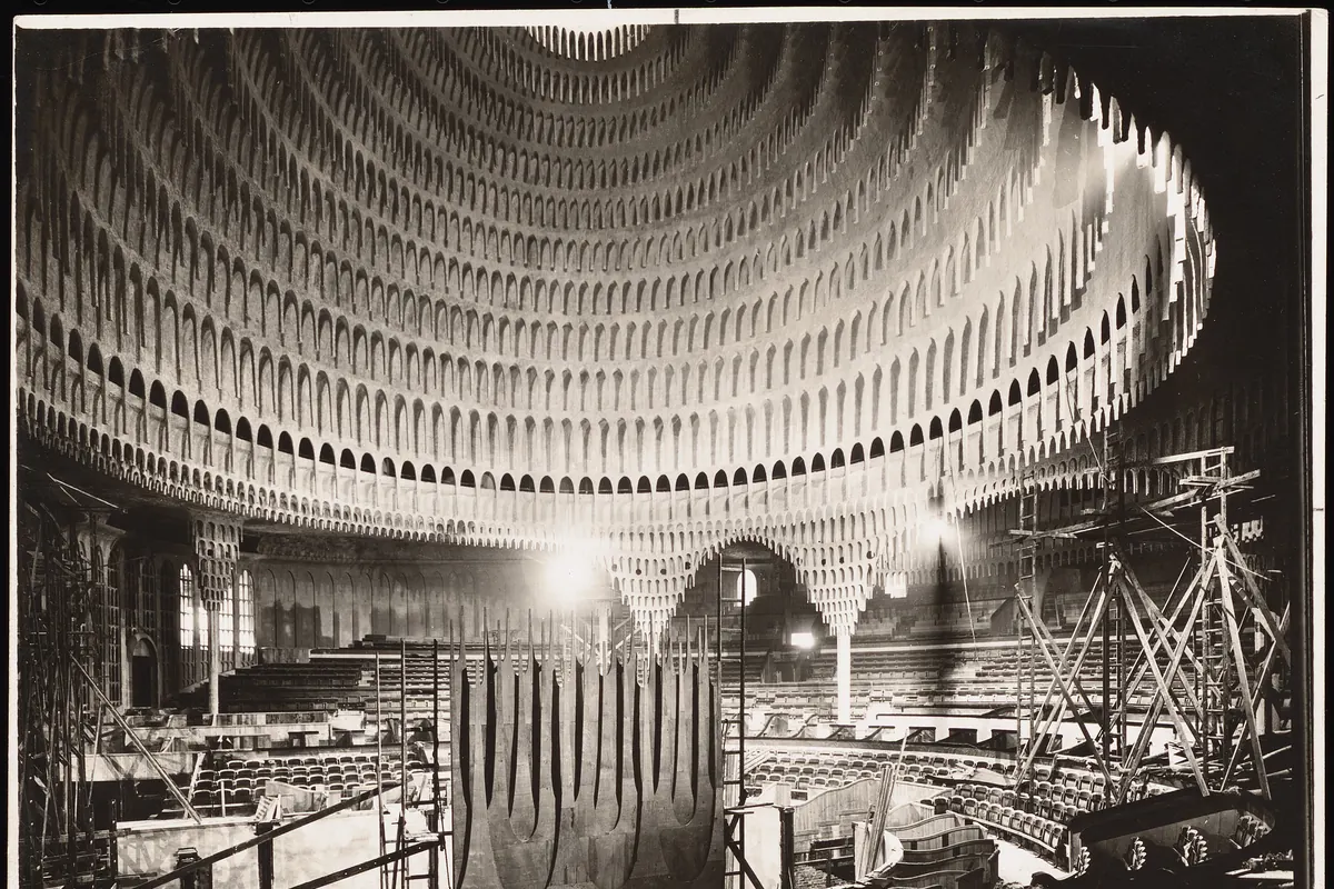 A circular theatre hall with plaster forms hanging from a domed ceiling over rows of seats, photographed in black and white