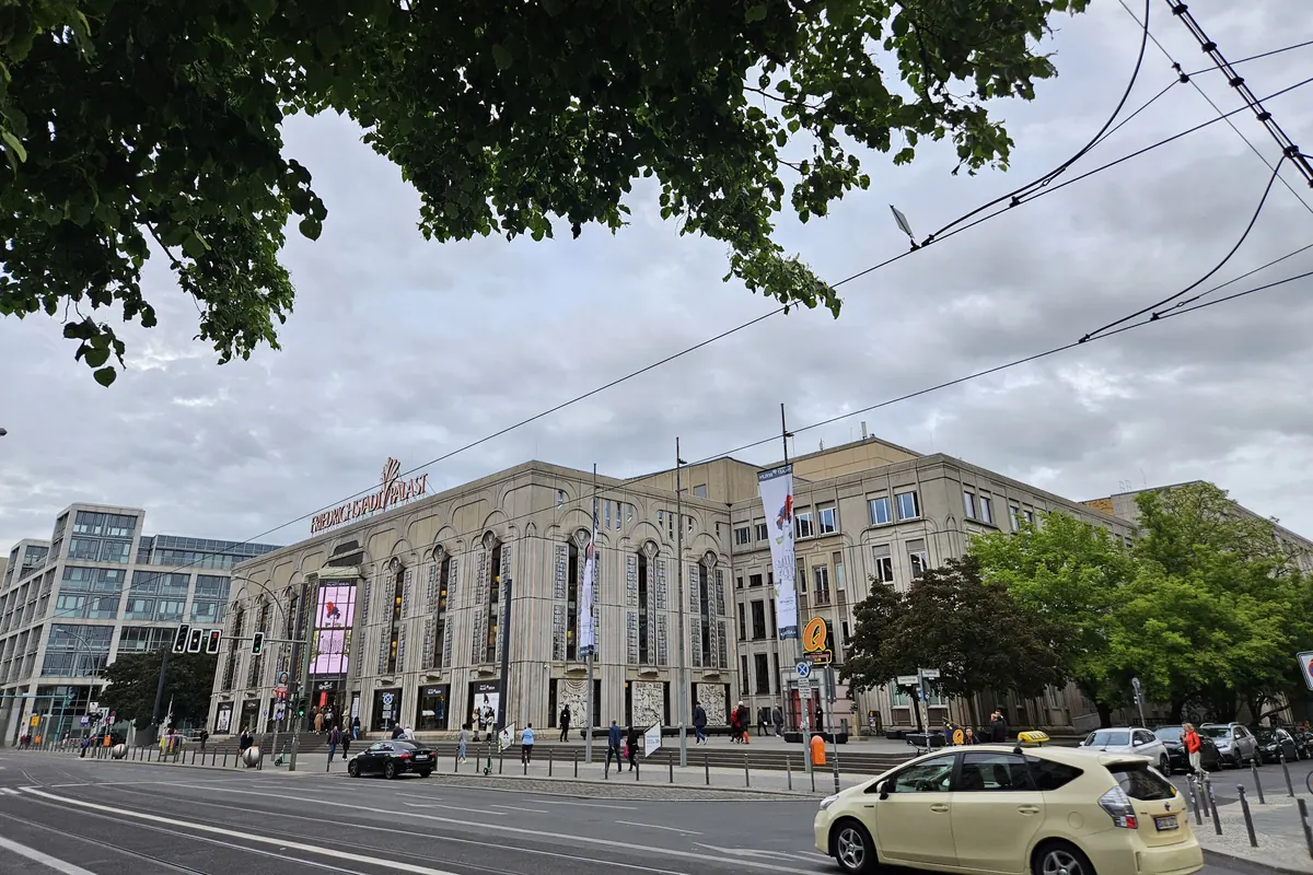 The stone front of a large theatre on a Berlin street, with tall arched windows, a glass entrance tower and a sign on the roof