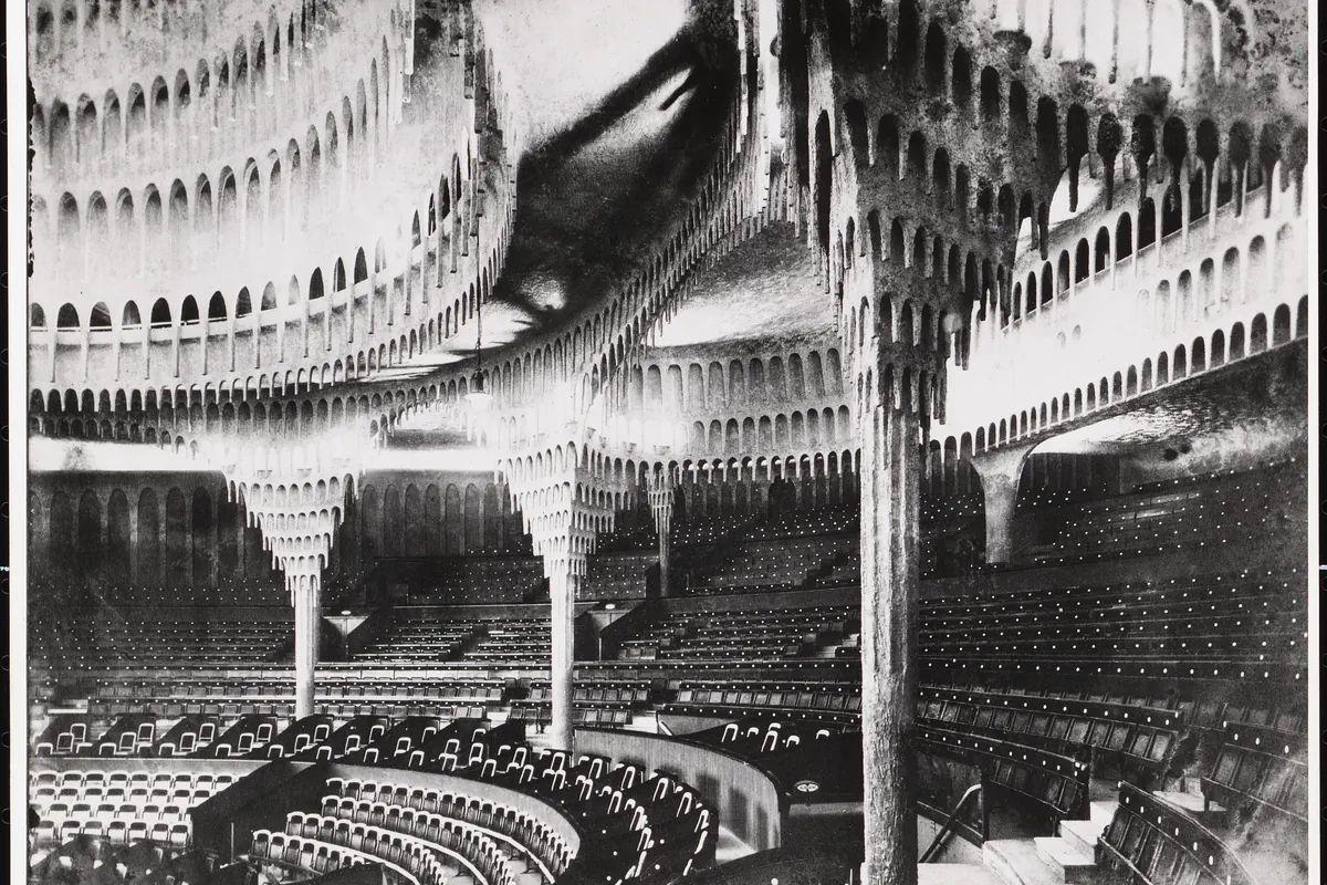A round theatre hall with tiered seating, plaster forms hanging from a domed ceiling and slender columns, photographed in black and white