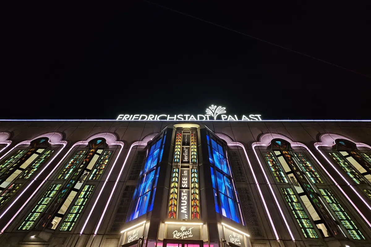 The front of a large theatre on a Berlin street at night, with a lit sign on the roof, tall arched windows glowing and people crossing the street