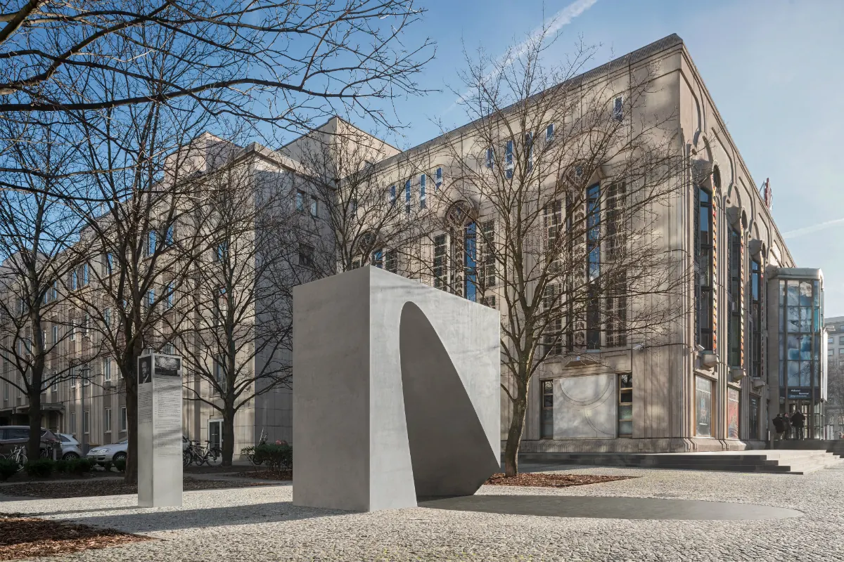 A pale concrete memorial block with a curved hollow, set among trees in front of the Palast, commemorating its founders
