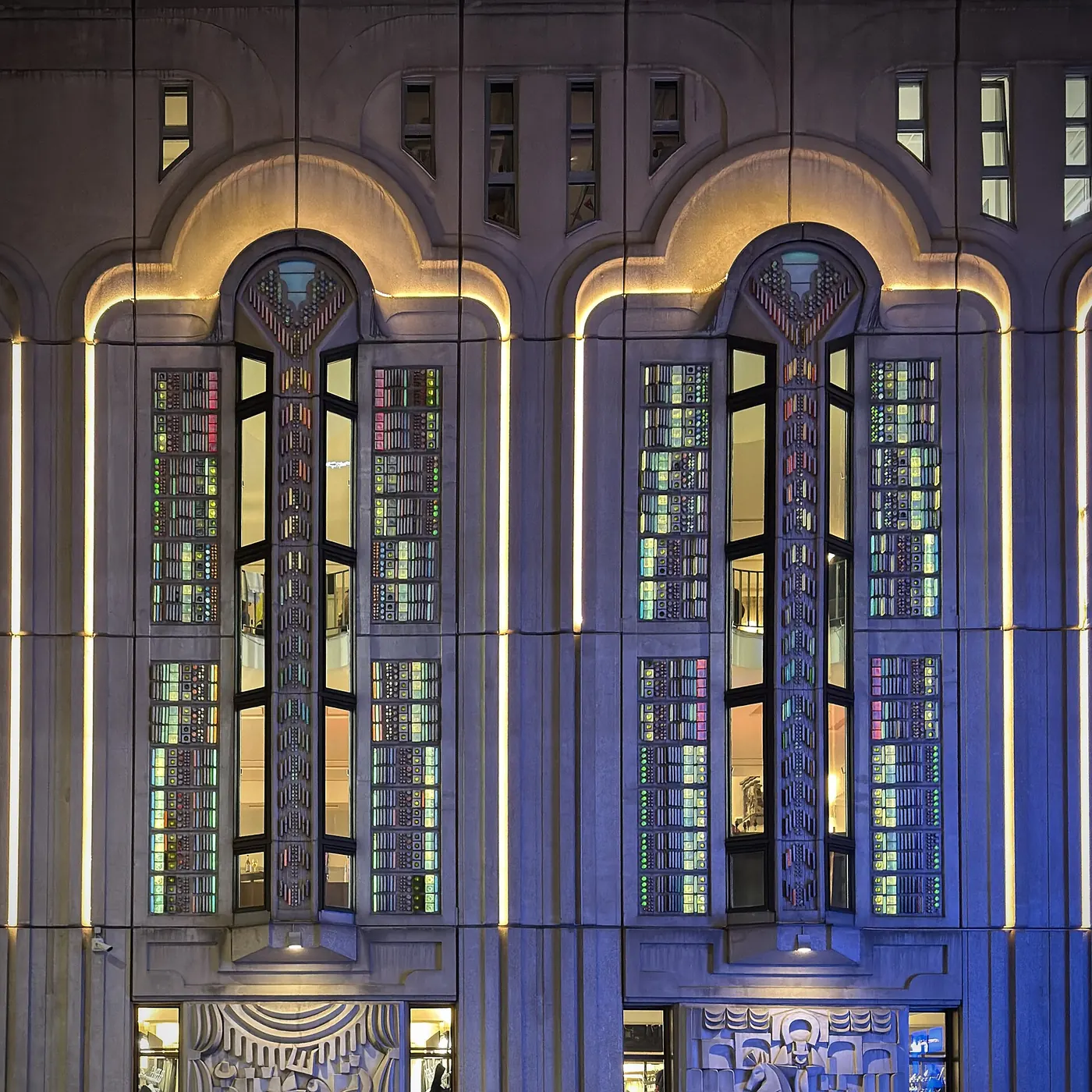 Two arched windows on the facade of the Palast at night, lit in blue and gold, with stained-glass panels set into the stone
